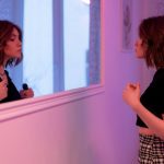 A brunette woman with bob haircut gazes at her reflection in the mirror, indoors with soft lighting.